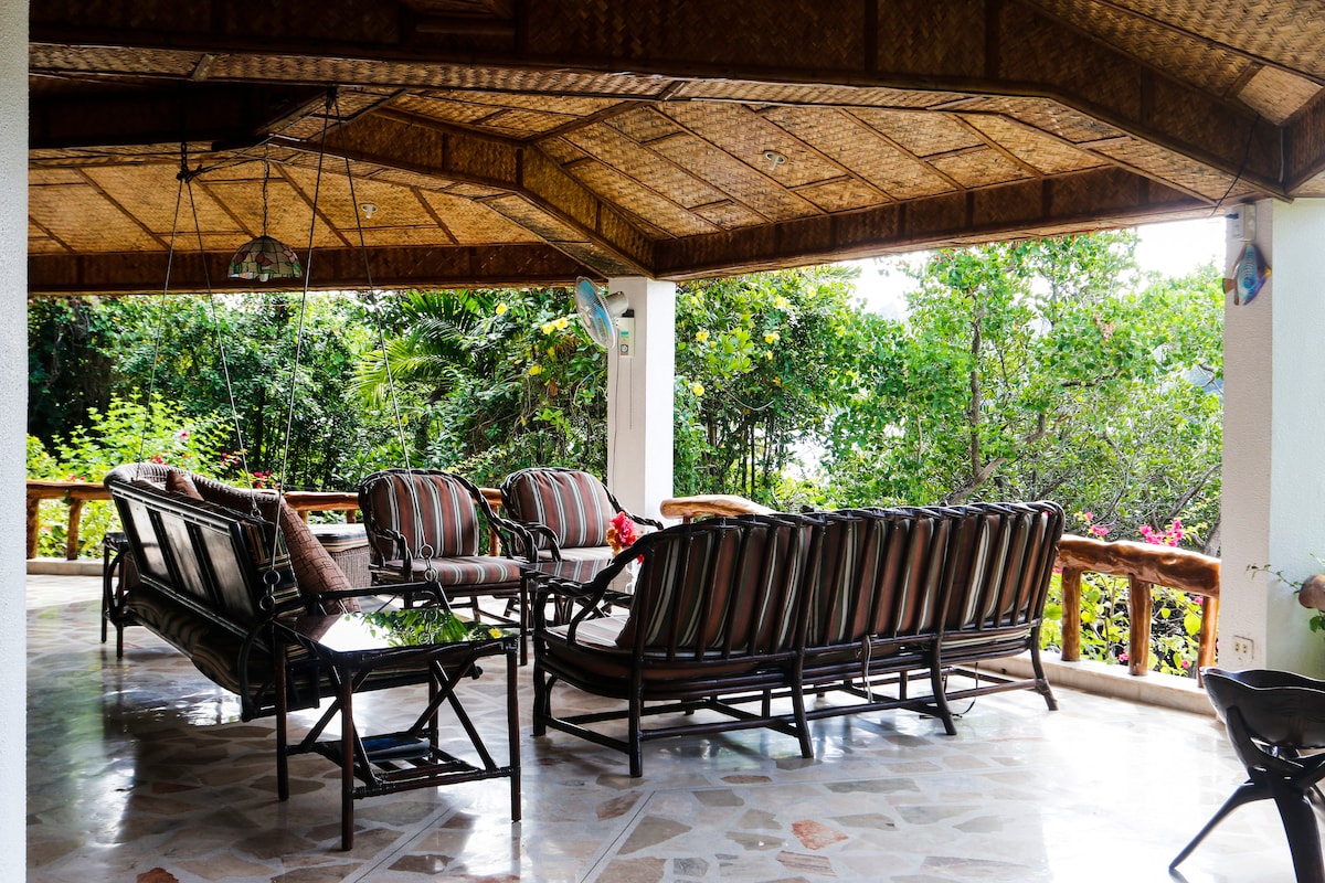 A spacious terrace is furnished with comfortable rattan lounge chairs and a glass-topped table. The area features a bamboo ceiling and is surrounded by lush greenery, providing a serene atmosphere with views of the vibrant landscape.