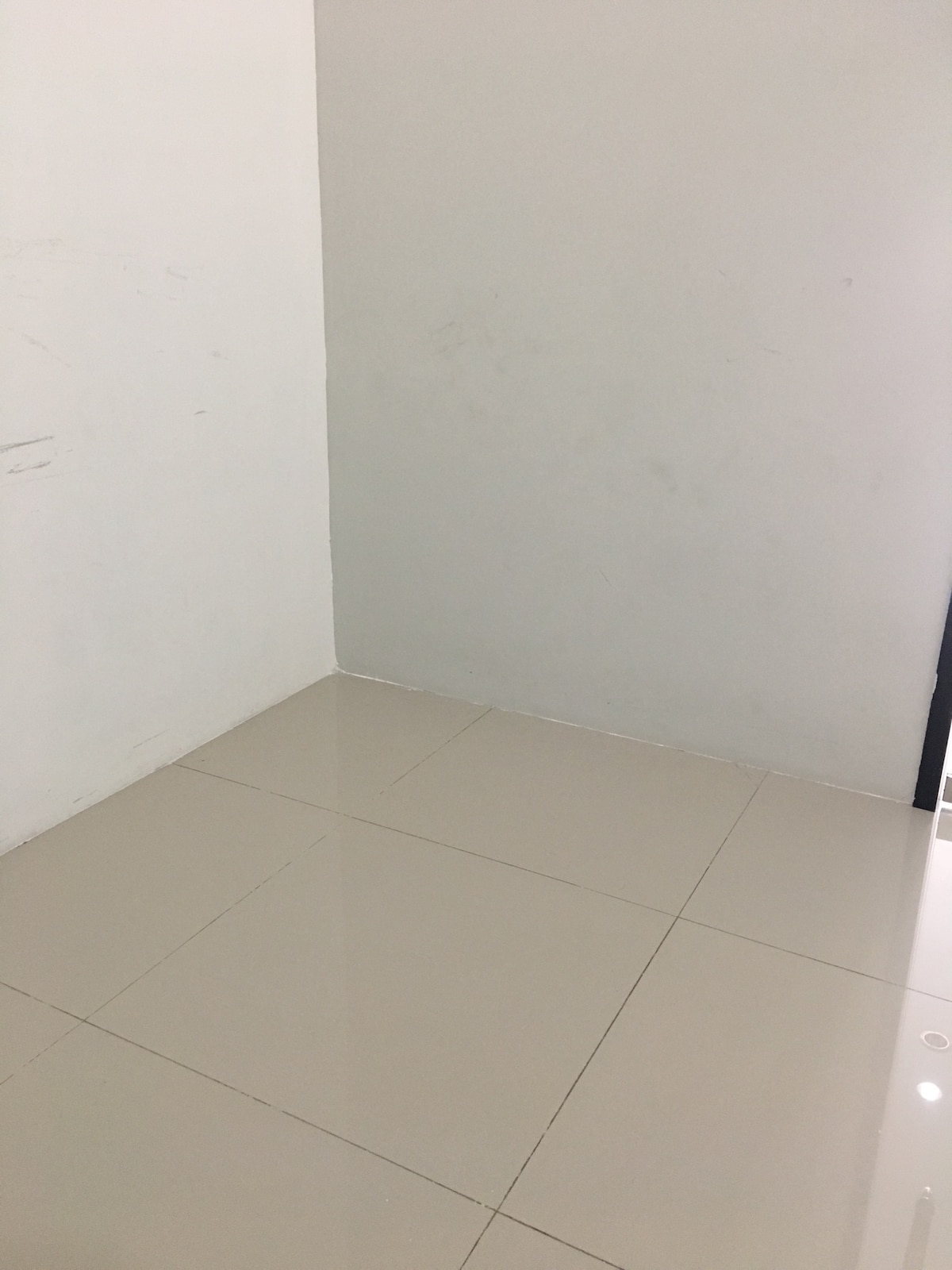 A minimal corner of a room is depicted, featuring smooth, glossy tiles that reflect light. The walls are painted in a light color, contributing to a clean and open feel. The image captures the simplicity of the space without any furnishings or decor.