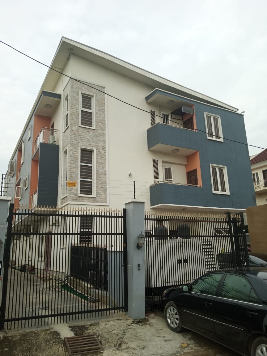 A three-story apartment building is shown, featuring a mix of stone and painted exterior walls. Multiple balconies are visible, framed by large windows. A gated entrance is present, with a black car parked nearby, and the surrounding area reflects urban living.