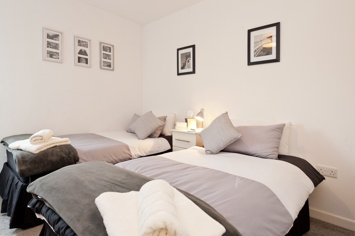 A guest bedroom is presented with two twin beds fitted with neutral-colored linens. Soft gray and white pillows are placed on each bed. A nightstand beside each bed holds a lamp, and black and white framed photographs decorate the wall.