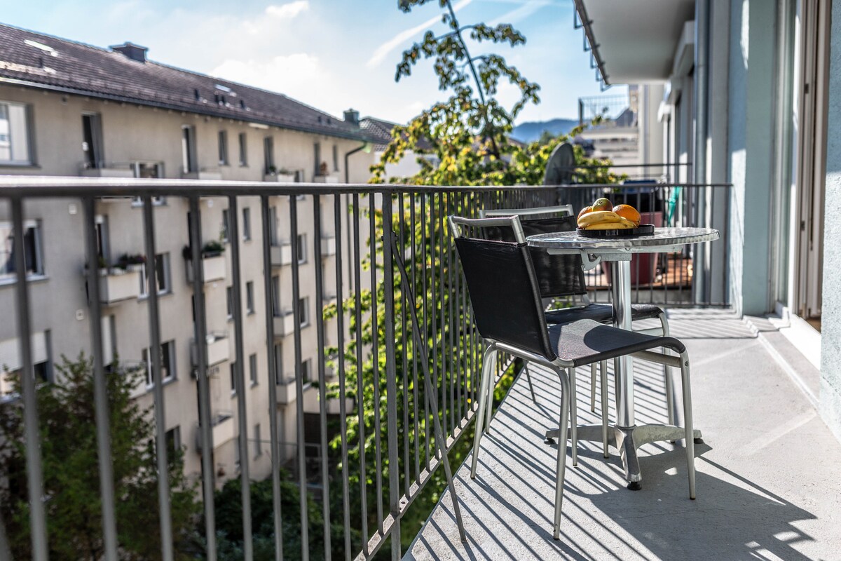 A balcony with a small table and two chairs overlooks a courtyard. Fresh fruit is displayed on the table, with sunlight casting shadows on the floor. The surrounding buildings are partially visible, framed by trees that enhance the outdoor space.