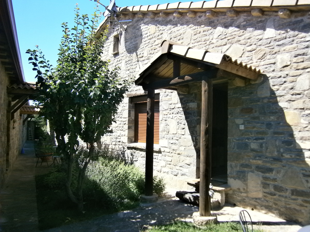 The exterior of a stone apartment is visible, featuring a wooden awning above the entrance. A small tree is positioned nearby, and the surroundings include well-maintained greenery. The building is complemented by a charming roof with tiles, indicative of traditional design.