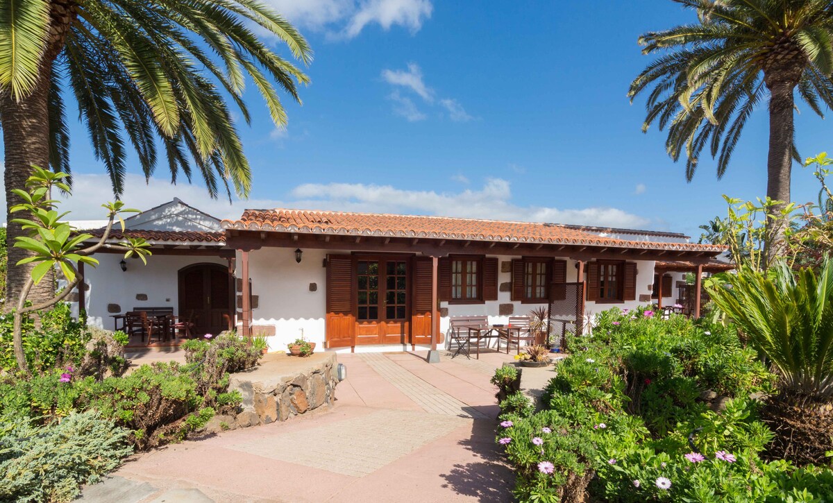 A charming rustic building is framed by palm trees, featuring a terracotta roof and wooden doors. The entrance is adorned with a pathway leading through lush gardens with vibrant flowers, inviting guests to explore the serene outdoor space.