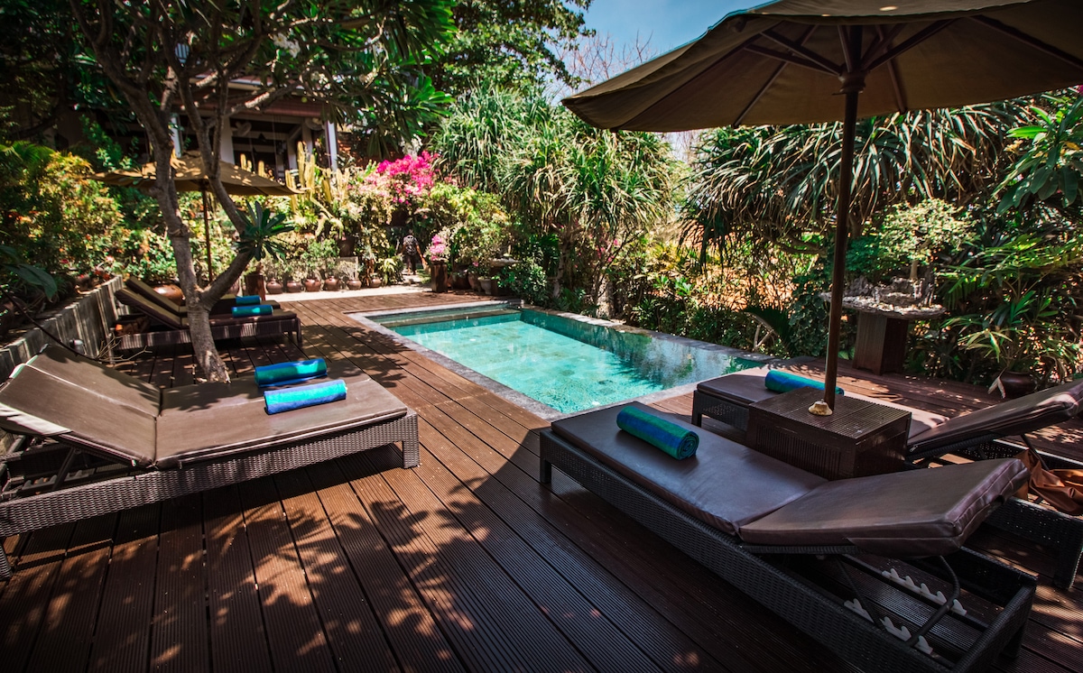 A serene outdoor space features a private swimming pool surrounded by lush foliage. Comfortable lounge chairs with rolled towels are positioned around the pool area, inviting relaxation. A large umbrella provides shade, complementing the natural beauty of the garden setting.