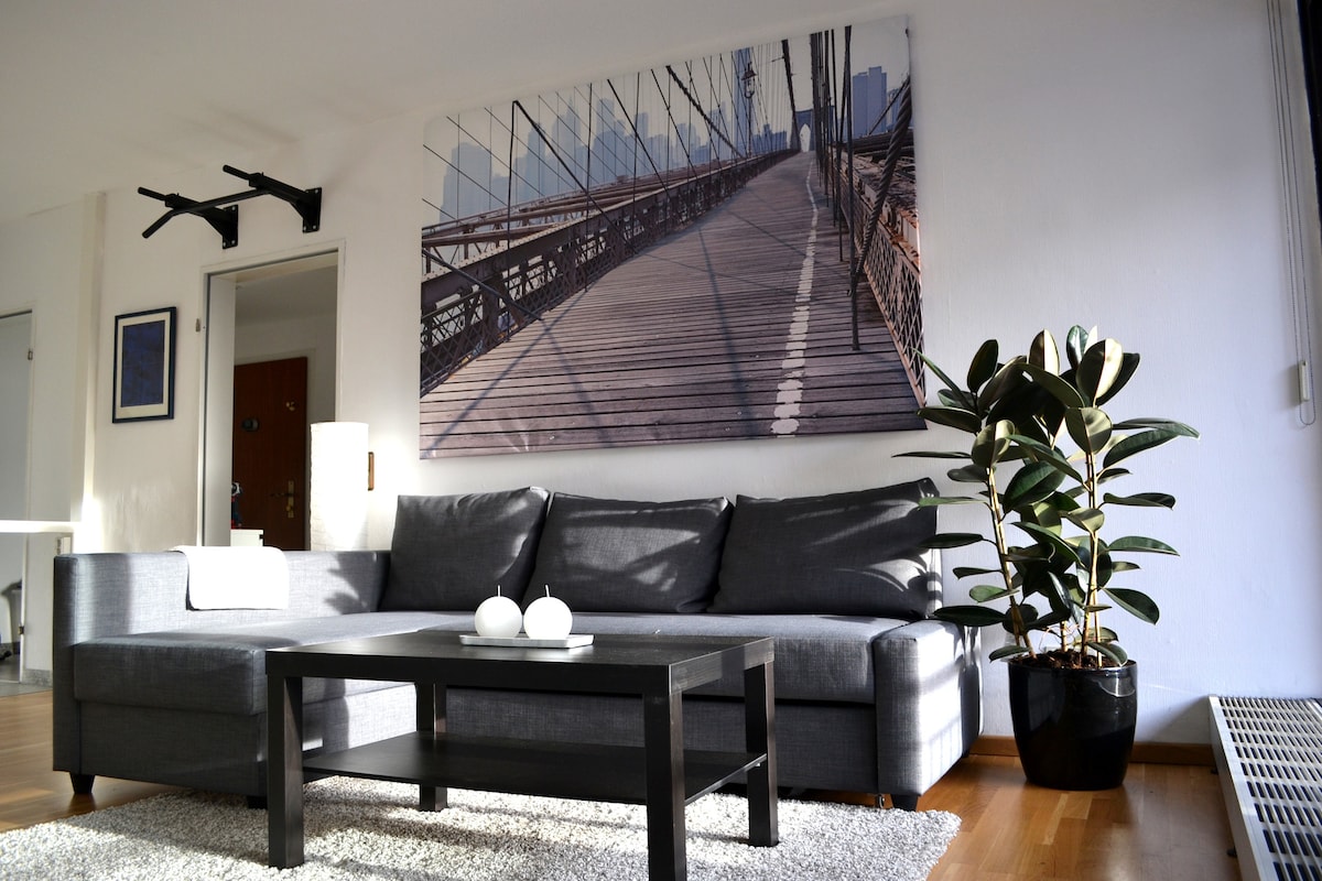 A modern living area features a spacious gray sofa paired with a dark coffee table, positioned on a light rug. A large wall art piece of a bridge adds visual interest, while a potted plant provides a touch of greenery. Natural light fills the space through nearby windows.