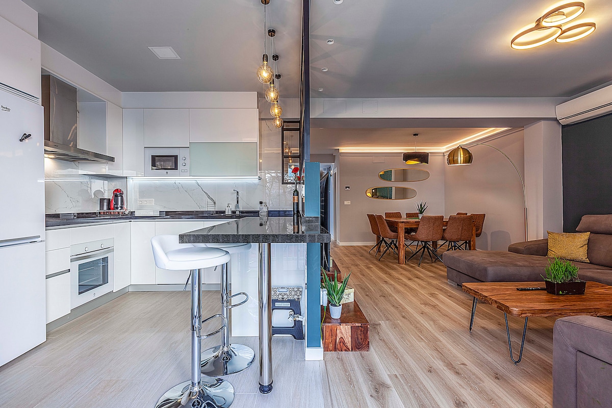 An open-plan living space combines a modern kitchen with a dining area. Sleek white cabinetry contrasts with dark countertops. A wooden dining table surrounded by beige chairs is visible, along with a comfortable seating area featuring a grey couch and wooden coffee table. Soft lighting enhances the atmosphere.