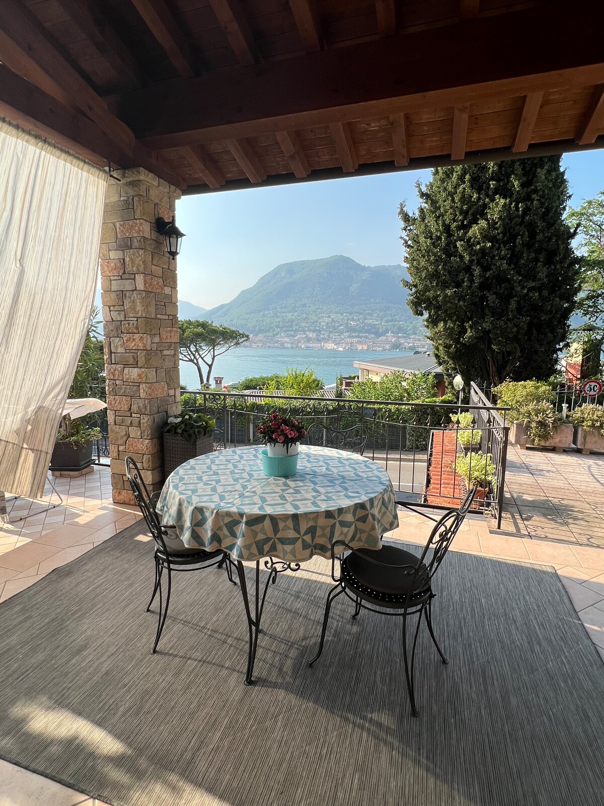 A covered terrace features a round table with a patterned tablecloth surrounded by two black chairs. Lush greenery and a view of the mountains and lake can be seen in the background, contributing to a serene outdoor dining experience.