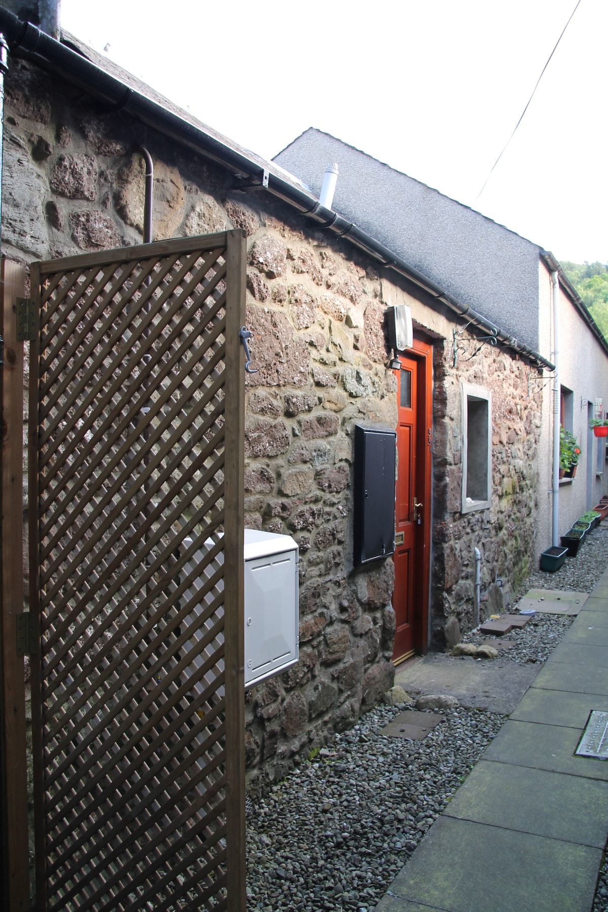 A charming stone exterior features a welcoming red door set within a narrow lane. The pathway is lined with small stones, and a latticed wooden gate provides entrance. The neighboring structures are visible along the sides, enhancing the quaint character of the area.