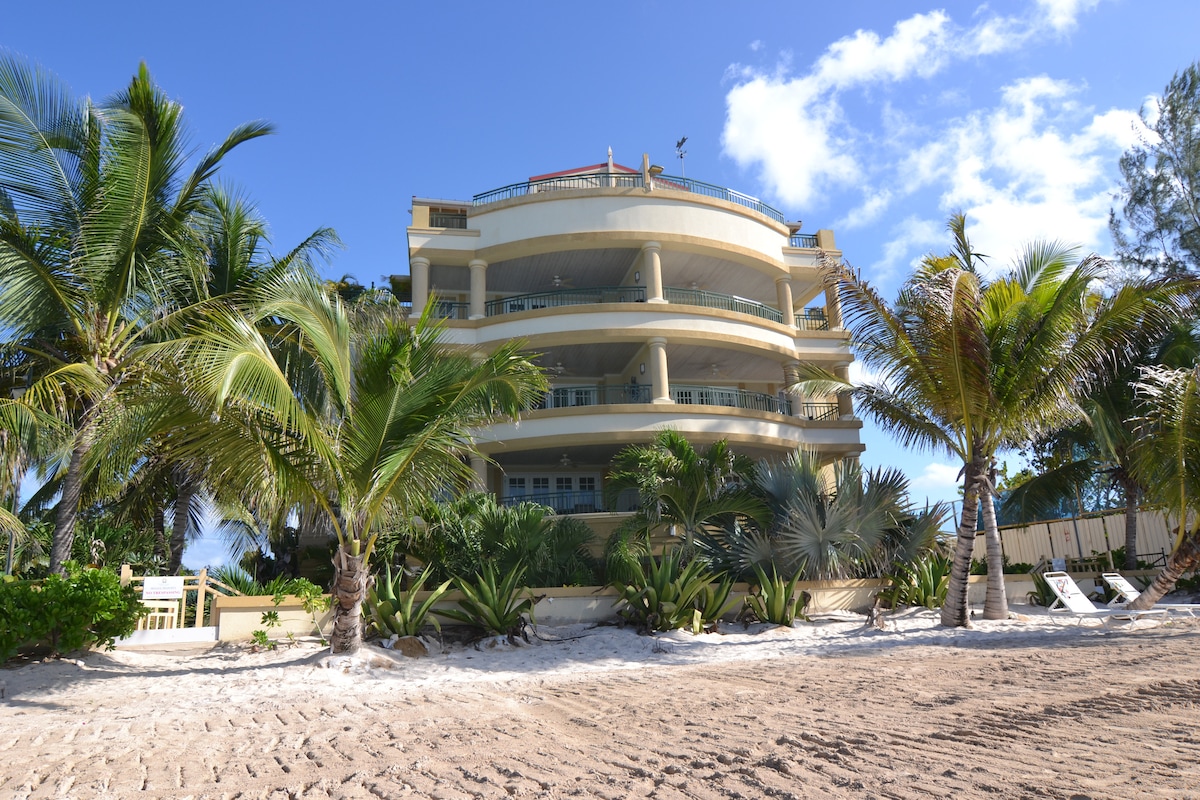 A three-story building is set amidst lush palm trees and coastal vegetation. The structure features multiple rounded balconies with ample outdoor seating. The sandy beach is visible nearby, inviting guests to enjoy the tranquil environment and the ocean's proximity.