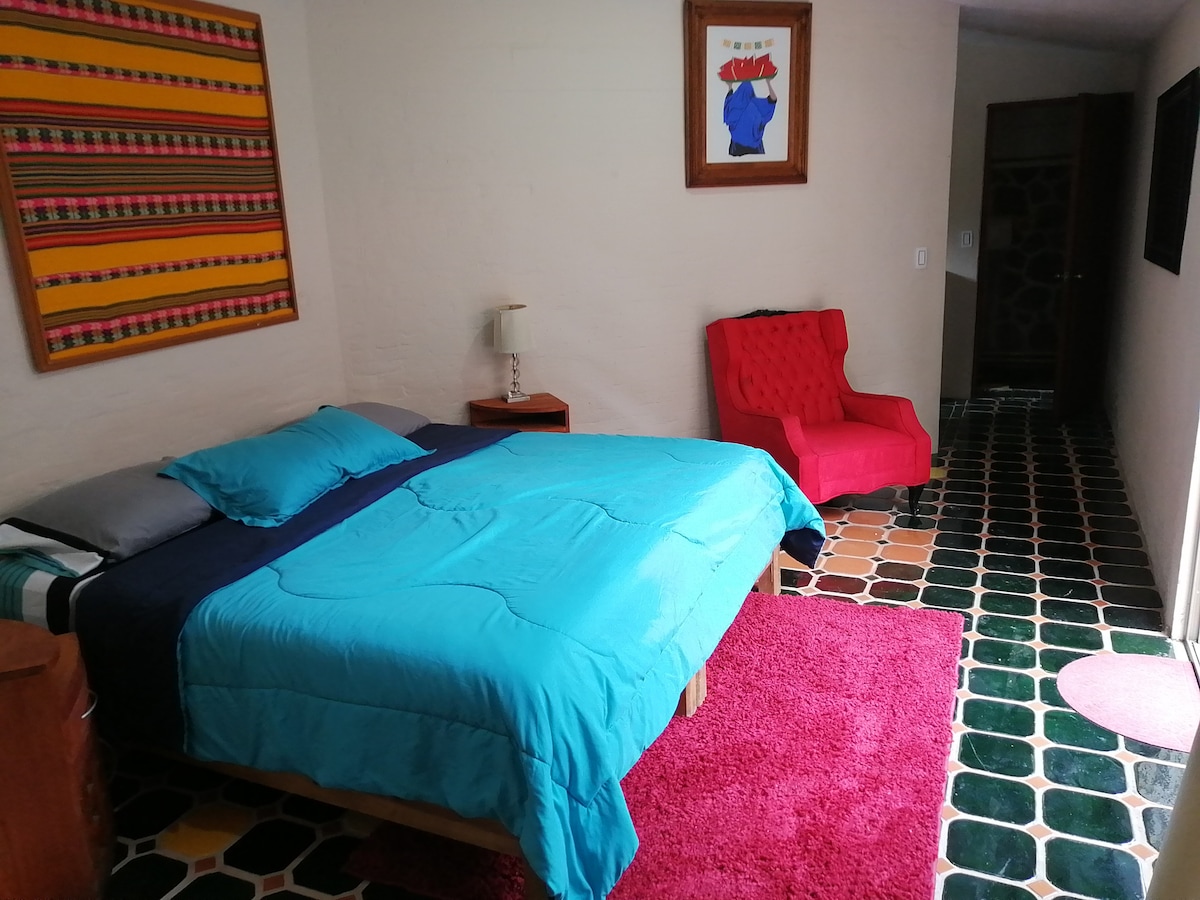 A spacious sleeping area is featured with a large bed covered in a vibrant blue duvet. A comfortable red chair adds a pop of color, and a colorful woven textile adorns the wall. The floor displays a unique tile pattern, contributing to the room's overall charm.
