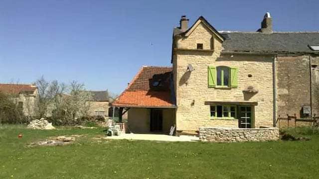 The exterior of a rustic stone house is shown, featuring a red-tiled roof and green shutters. A spacious lawn surrounds the home, with trees and farm buildings visible in the background. A small stone wall lines the patio area, providing a serene garden space.