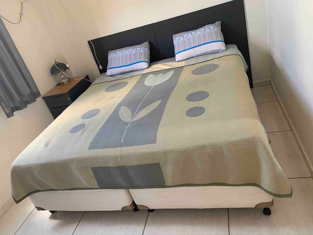 A spacious bed is positioned against a light-colored wall, adorned with a simple patterned bedspread featuring floral designs. Two pillows provide comfortable support. A bedside lamp and a dark nightstand are visible on one side of the bed, complementing the neutral decor.