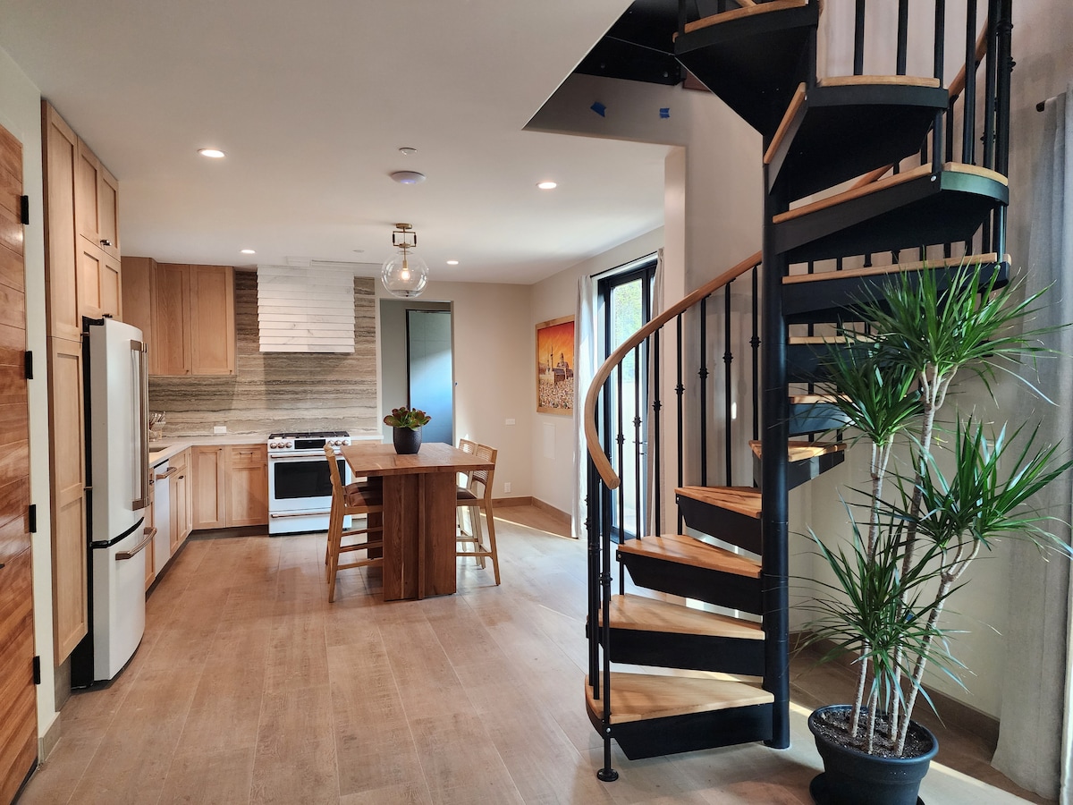 The open kitchen area features modern appliances and wood cabinetry, complemented by a dining table that seats four. A spiral staircase with wooden steps leads to the upper floor. Natural light illuminates the space through large windows, with potted plants adding a touch of greenery.