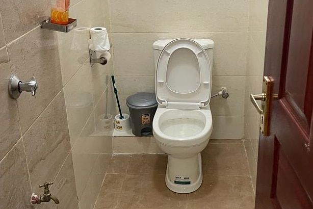 A clean bathroom is depicted, featuring a white toilet positioned against a tiled wall. Essential items such as toilet paper and a small trash bin are visible. A faucet is mounted on the left, with a wall-mounted dispenser above the toilet.