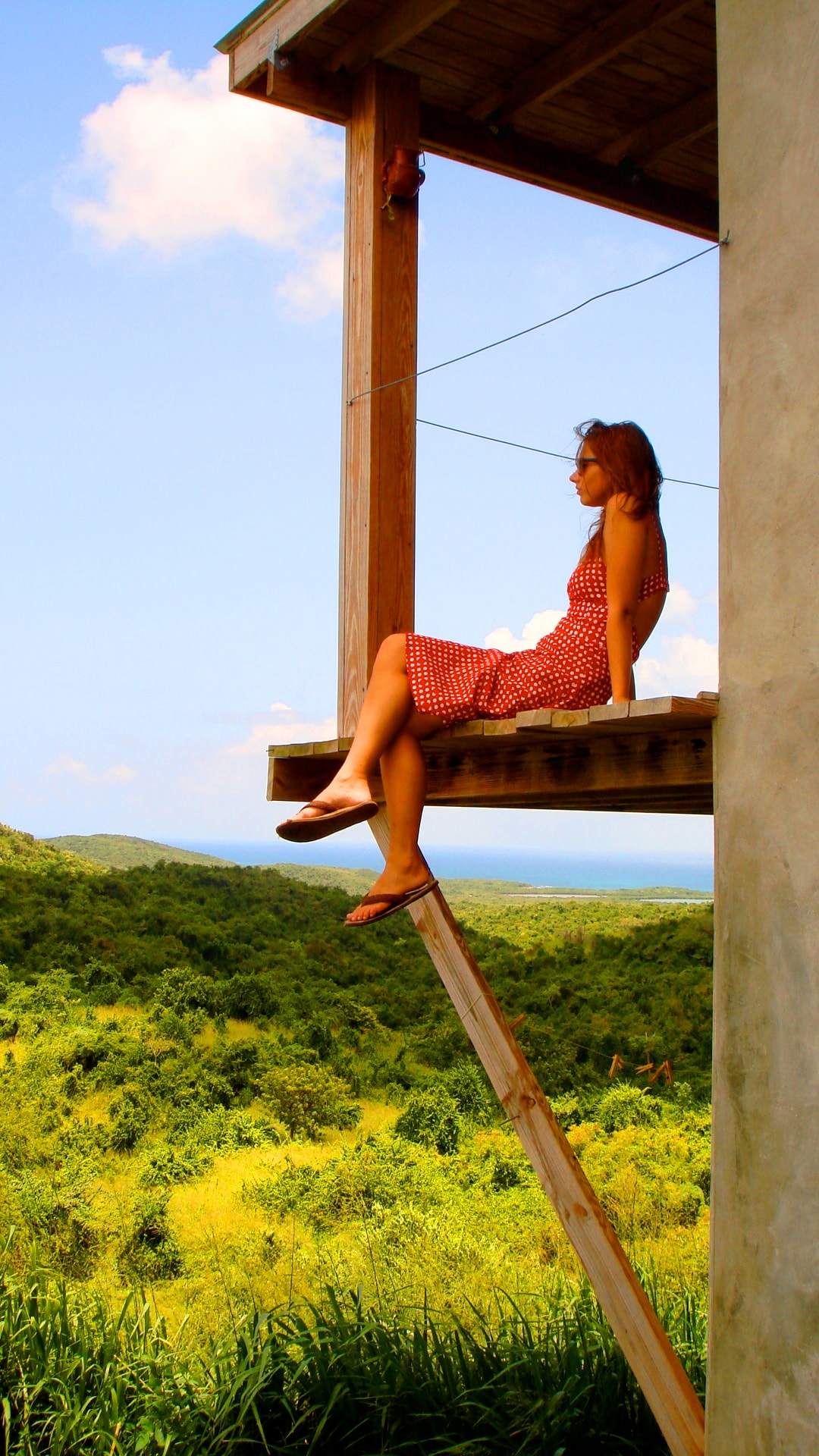 A wooden balcony extends from the two-story house, with a scenic view of lush greenery and the ocean visible in the distance. Natural light brightens the space under a clear blue sky, creating a serene and inviting outdoor setting.