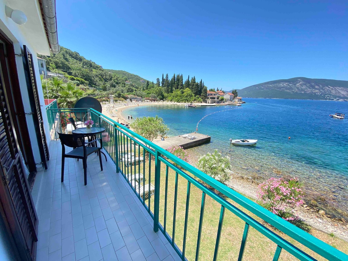 A balcony overlooks a tranquil bay with clear blue waters. Green shrubbery and trees frame the view, while boats can be seen in the distance. The area features a small beach, with a path leading to the water. Outdoor seating is arranged for guest use.