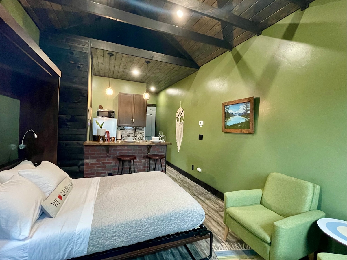 A spacious studio room is highlighted by vaulted ceilings with wood beams. A full-size Murphy bed is visible beside a reading chair, and a kitchenette featuring a breakfast bar is seen in the background. Soft green walls and natural light enhance the cozy atmosphere.