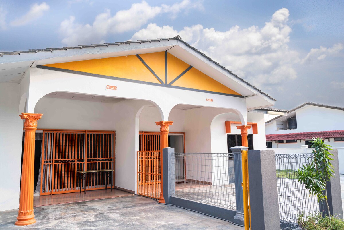 The exterior entrance features a bright orange and white color scheme, highlighted by tall columns. A covered area allows for sheltered entry. The space is surrounded by a wrought iron fence, offering a secure environment, with a clear view of the paved path leading to the front door.