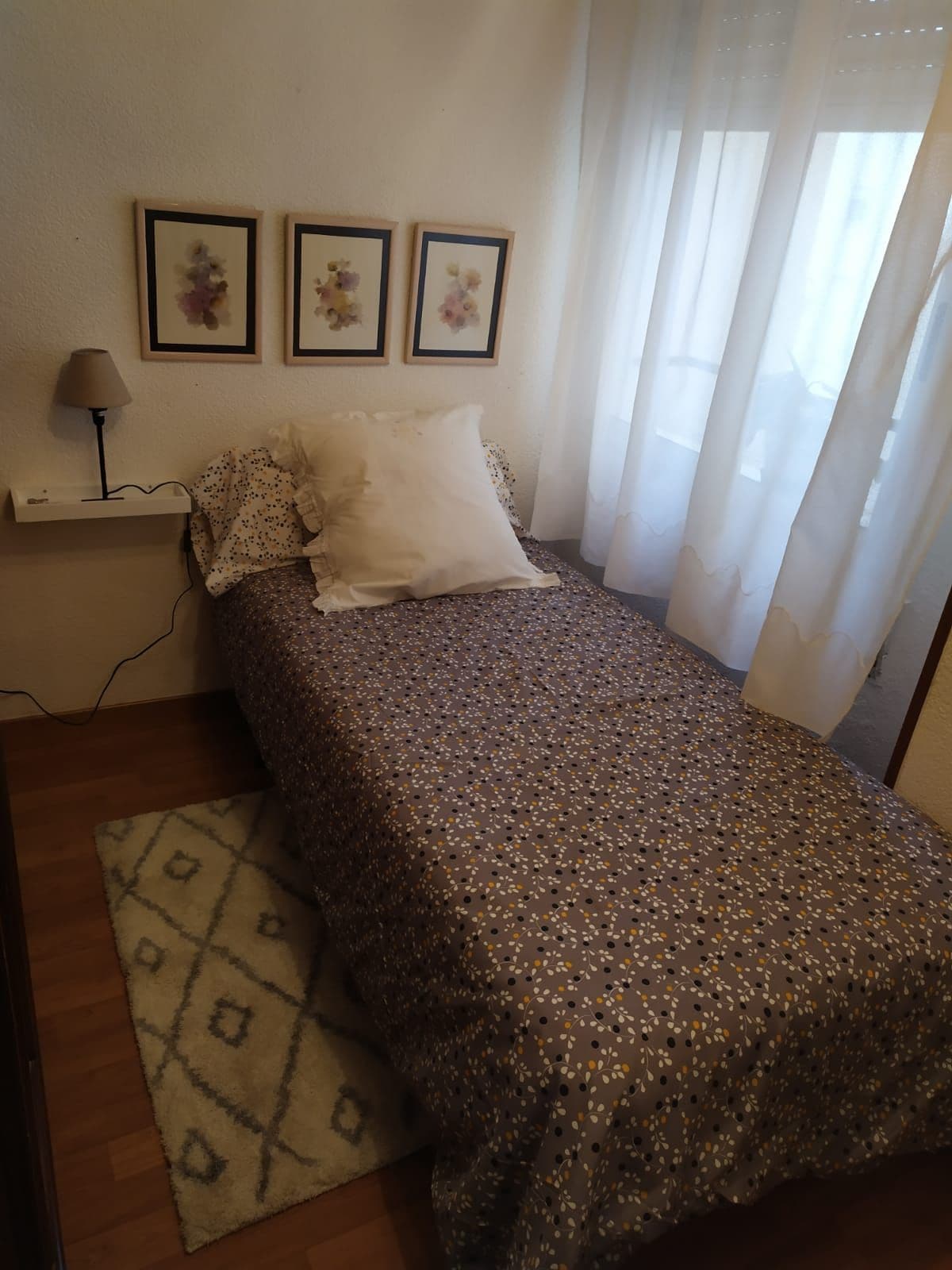 A compact bedroom features a single bed dressed in a decorative coverlet with a white and floral pattern. Three framed floral prints adorn the wall above the bed. Natural light filters through a sheer curtain, illuminating the space, while a small bedside lamp is positioned nearby.