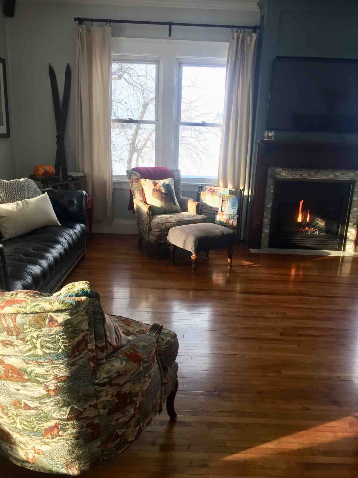 A warm living area features a plush, dark leather couch accompanied by two patterned armchairs. A cozy fireplace is situated near a large window, allowing natural light to illuminate the hardwood floor. Vintage decor elements add charm to the space.