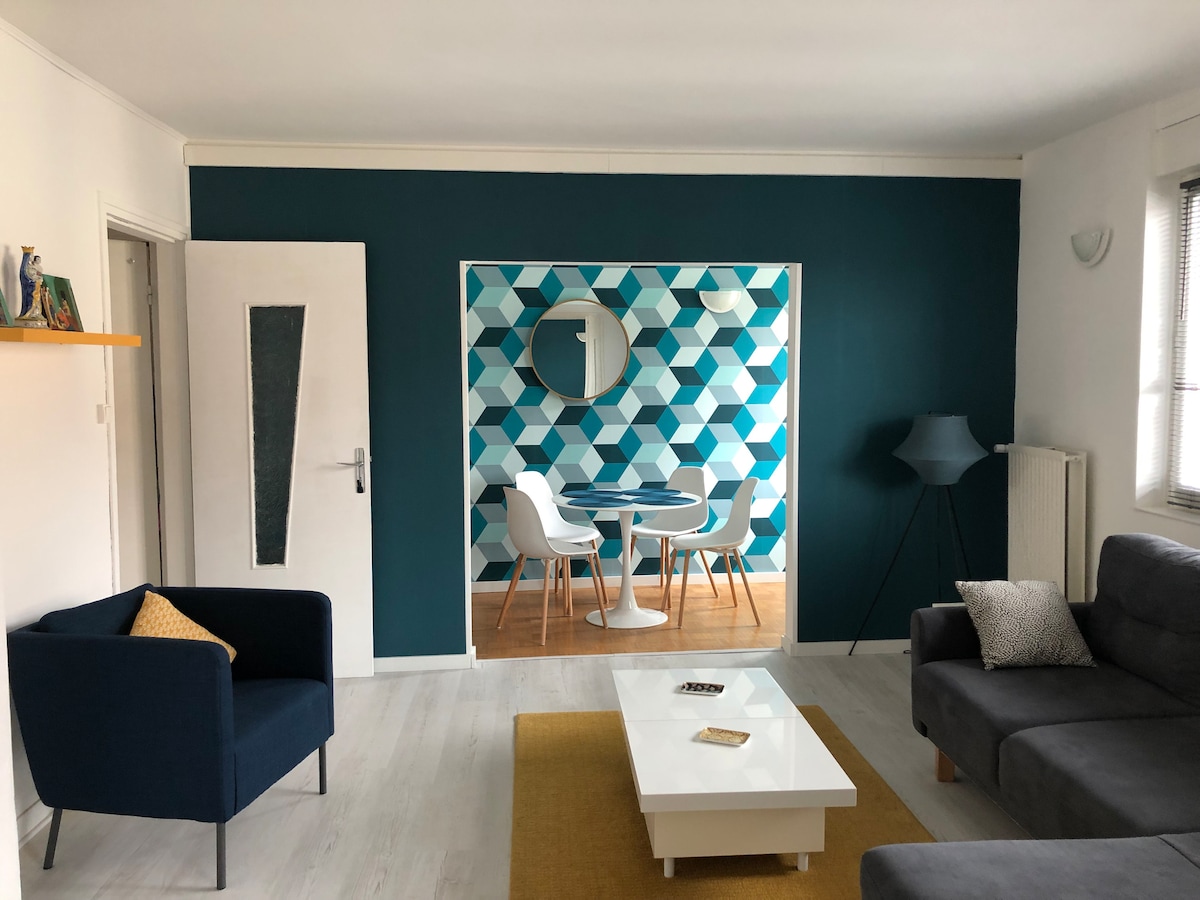 A contemporary living area features a mix of blue and white geometric wall patterns. A round dining table with white chairs sits in the background, while a light gray sofa and dark blue armchair, accompanied by a side table, create a comfortable seating space.
