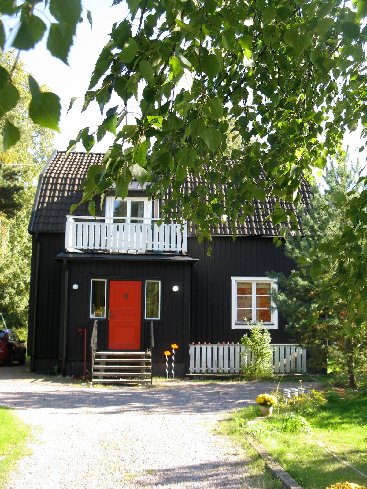 A two-story, dark wooden house is framed by lush green trees. A bright red front door is highlighted, with white railings bordering the upper balcony. A pebble driveway leads to the entrance, surrounded by carefully placed plants and flowers.