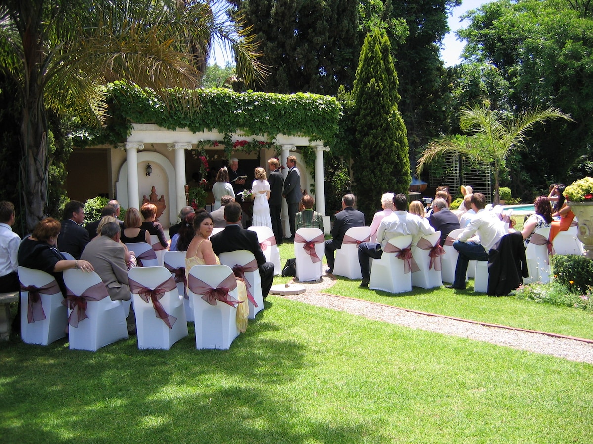An outdoor wedding ceremony takes place in a verdant garden. Guests are seated on white chairs adorned with burgundy sashes, while a couple stands at an altar framed by greenery. Lush trees and colorful floral arrangements enhance the serene setting.