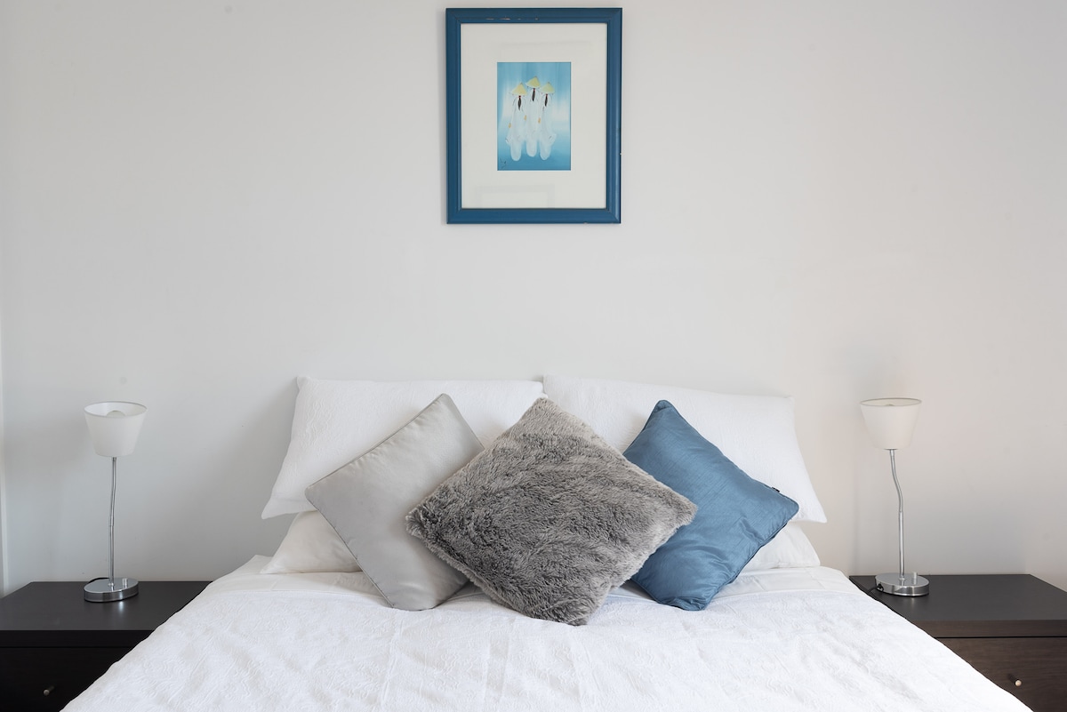A neatly arranged bed is presented with a variety of soft pillows in shades of gray and blue. A framed artwork hangs above the bed, complementing the light-colored walls. Two bedside lamps are positioned on dark wooden nightstands for additional illumination.