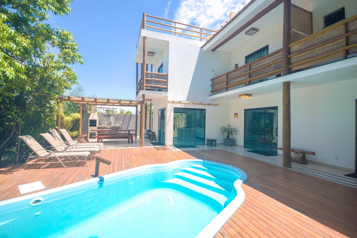 An outdoor area features a pool with steps for easy access, surrounded by wooden decking. Sun loungers are positioned nearby under a pergola, providing shaded seating. The property is enhanced by green foliage, contributing to a relaxed atmosphere.