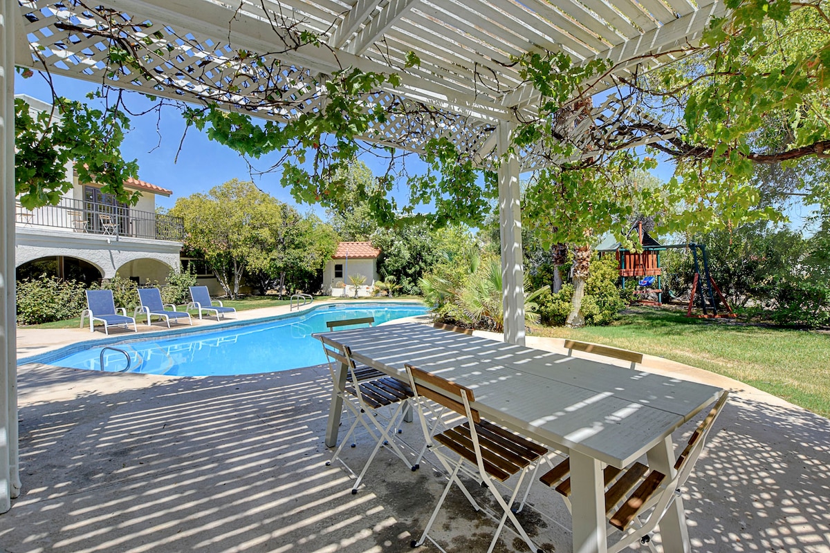 A shaded pergola overlooks a private pool, framed by lounge chairs beneath a clear blue sky. A dining table with wooden slats is positioned nearby, surrounded by lush greenery and tall trees, with an outdoor playset visible in the background.