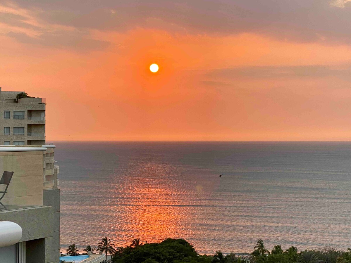 A serene sunset is captured over the ocean, with warm orange and pink hues reflecting off the water. The view includes a faint silhouette of distant palm trees, enhancing the peaceful atmosphere of the coastal landscape.