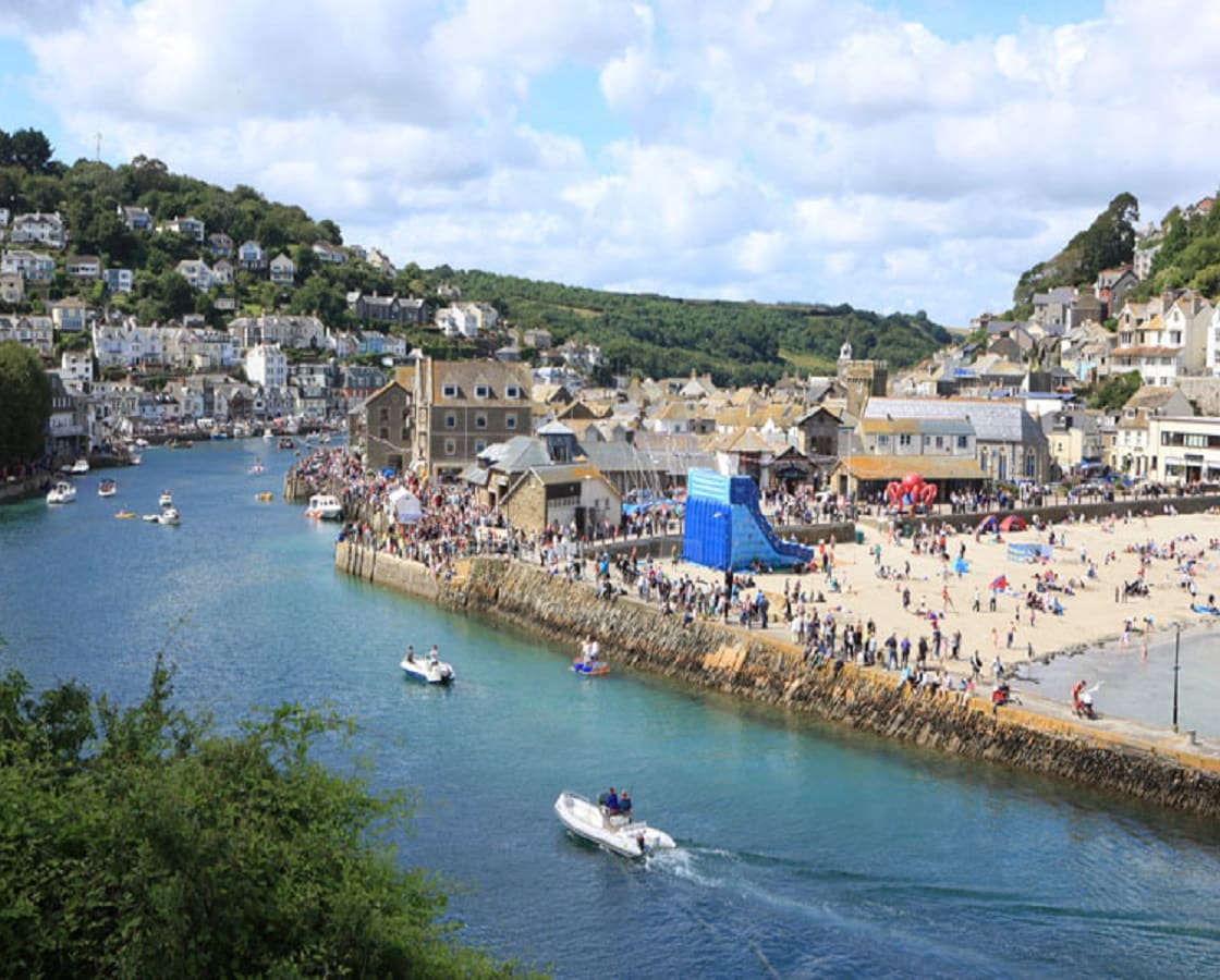 A seaside view captures the bustling harbor area, featuring a sandy beach lined with visitors. Small boats can be seen gliding through the vibrant blue water, while the lush green hillside and charming buildings provide a picturesque backdrop.