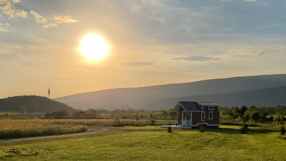 The tinyhouse is situated on open land, framed by rolling hills and a vibrant sunset sky. Warm sunlight casts a golden hue over the grassy landscape, creating a serene backdrop for the home.