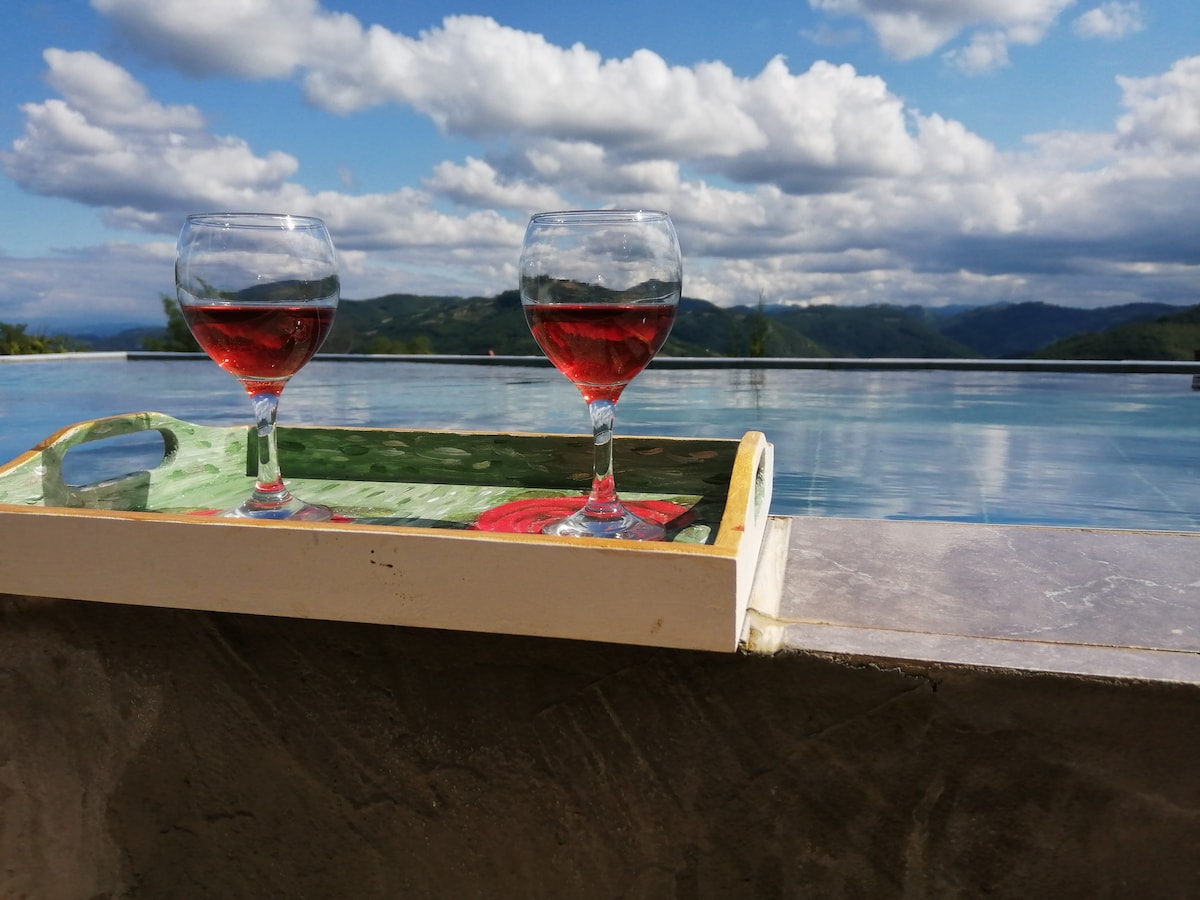 Two glasses of red wine are placed on a tray by the edge of an infinity pool. The tranquil water reflects the sky, with distant mountains visible in the background under a partly cloudy atmosphere.