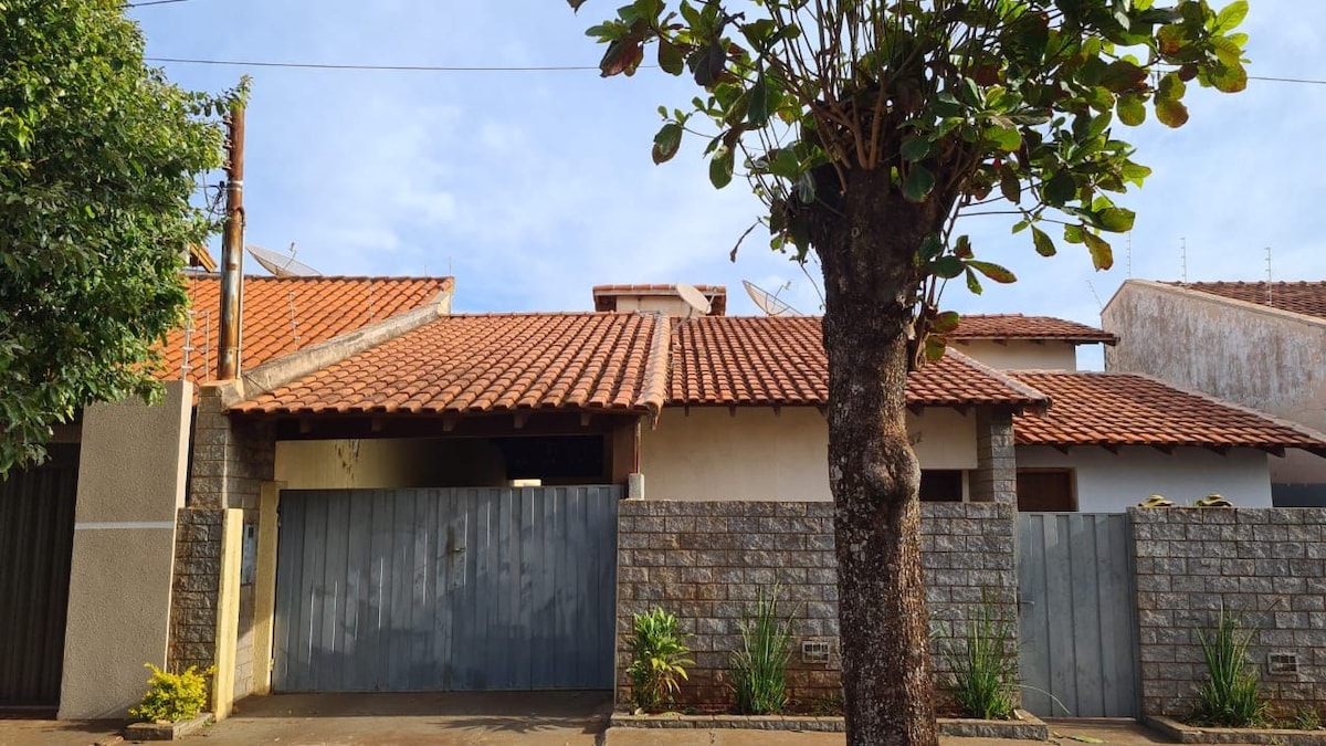 A charming house with a terracotta roof is framed by a lush tree and greenery. The entrance features a wooden gate in front of a stone wall, providing an inviting façade to the property.