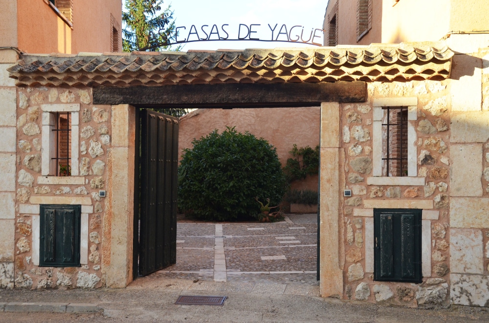 The entrance to the property is marked by a wooden gate framed in stone. An inviting pathway leads into a courtyard, surrounded by greenery. The structure is complemented by low stone walls and a sign that reads 'Casas de Yagué' above the entrance.