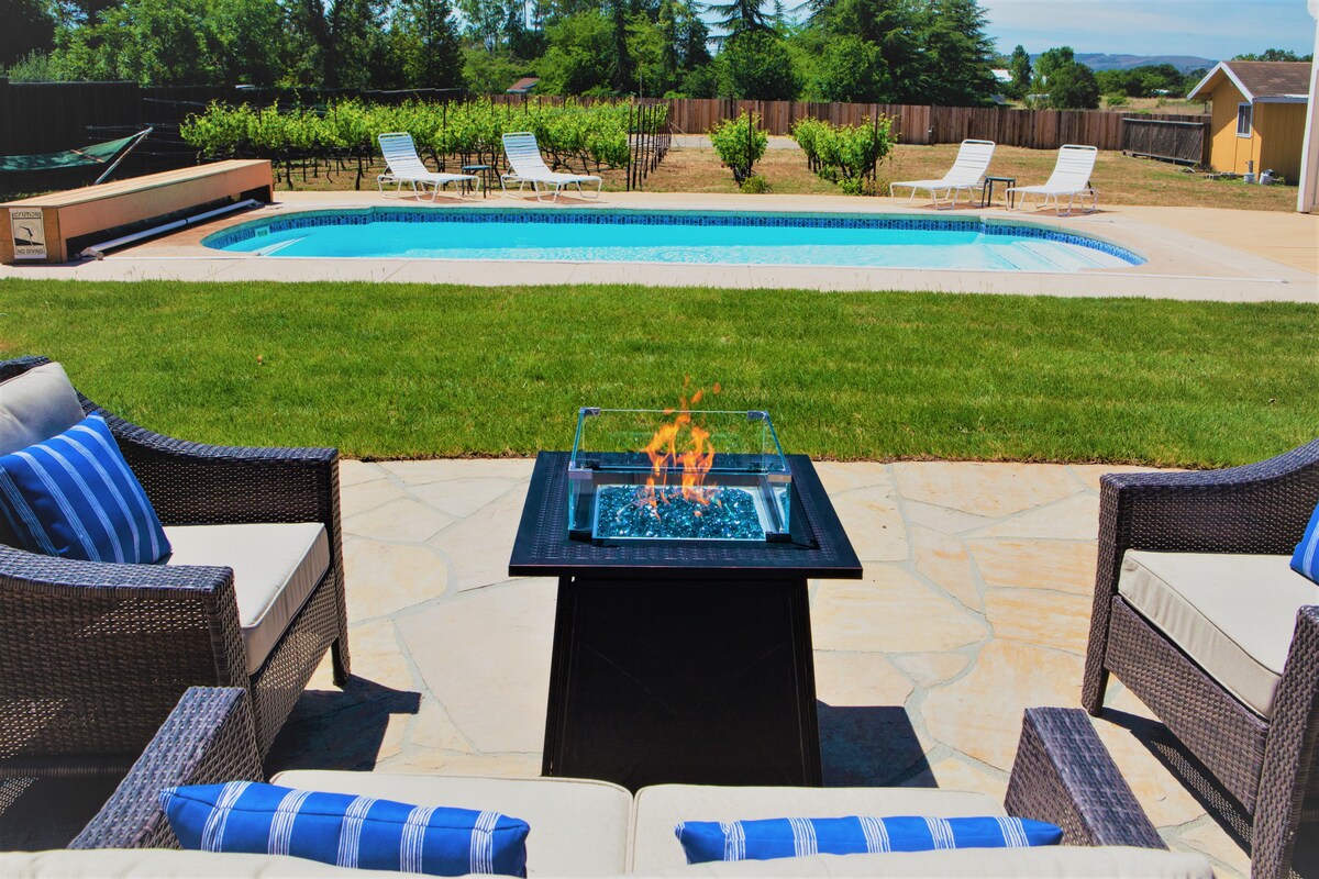 An outdoor seating area features plush couches with blue striped pillows surrounding a fire pit. In the background, a solar-heated pool reflects clear skies, while lounge chairs are positioned nearby on a landscaped lawn integrating views of adjoining vineyards.
