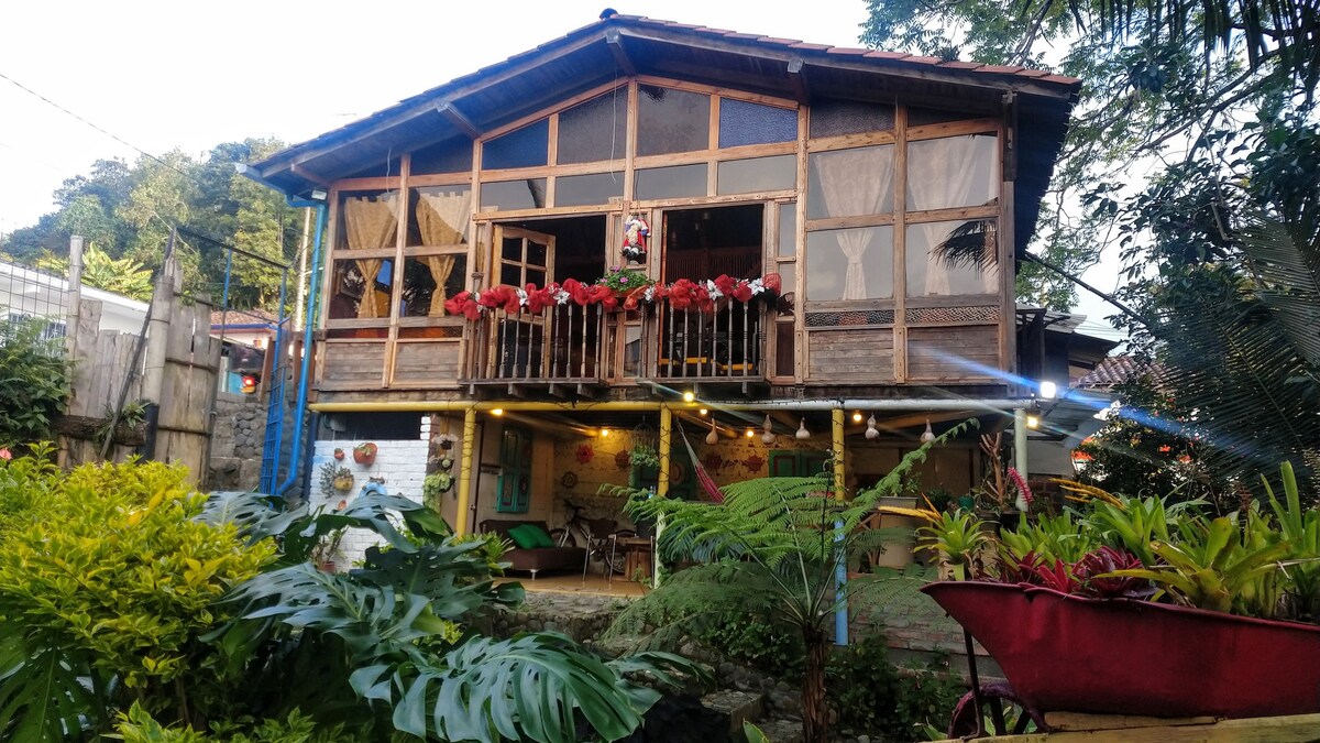 The charming cabin exhibits a wooden facade with large glass windows, allowing natural light to fill the space. Vibrant flowers are displayed on the balcony, complemented by lush greenery in the garden area below. A welcoming outdoor seating area can be seen, enhancing the overall ambiance.