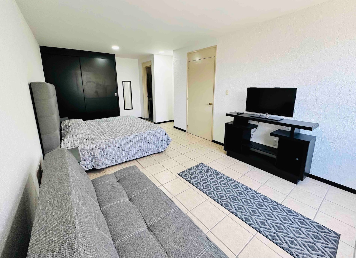 A spacious room is visible, featuring a large bed with a patterned coverlet and a modern flat-screen TV on a simple stand. A gray sofa bed provides additional seating. Light tile flooring enhances the open feel of the space, complemented by a patterned area rug.