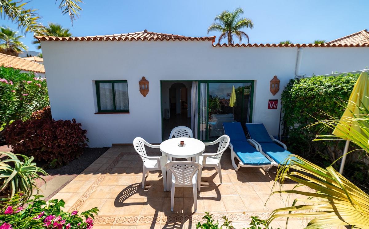 A charming outdoor patio features a round table surrounded by four white chairs, complemented by two lounge chairs. Lush greenery and potted plants provide a vibrant backdrop, with a view of the bungalow's access door and large glass windows reflecting the sunny sky.