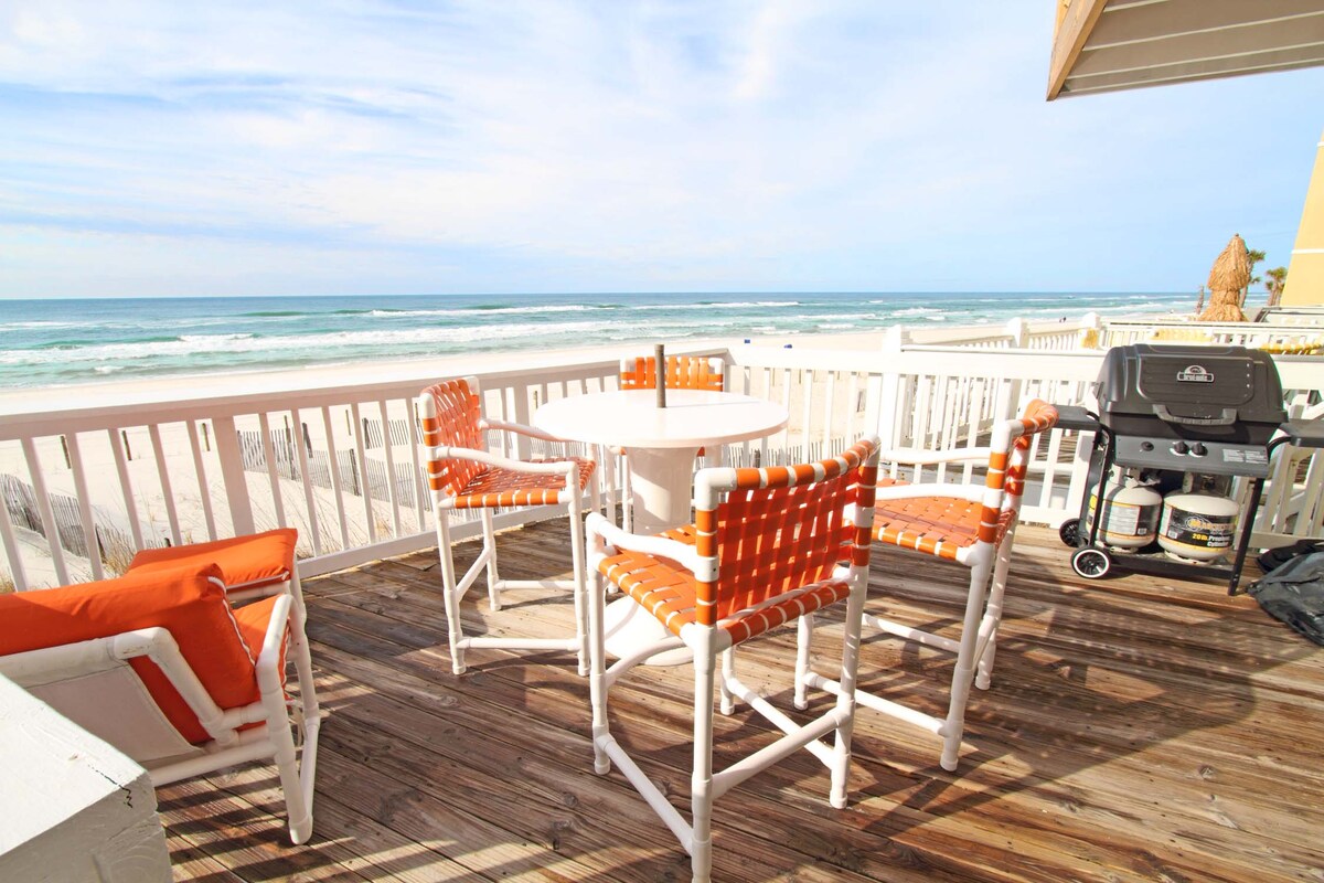 An outdoor patio space features a round table surrounded by six white chairs with orange woven backs. A grill stands nearby, offering a view of the ocean and waves, while wooden decking provides a warm and inviting surface.