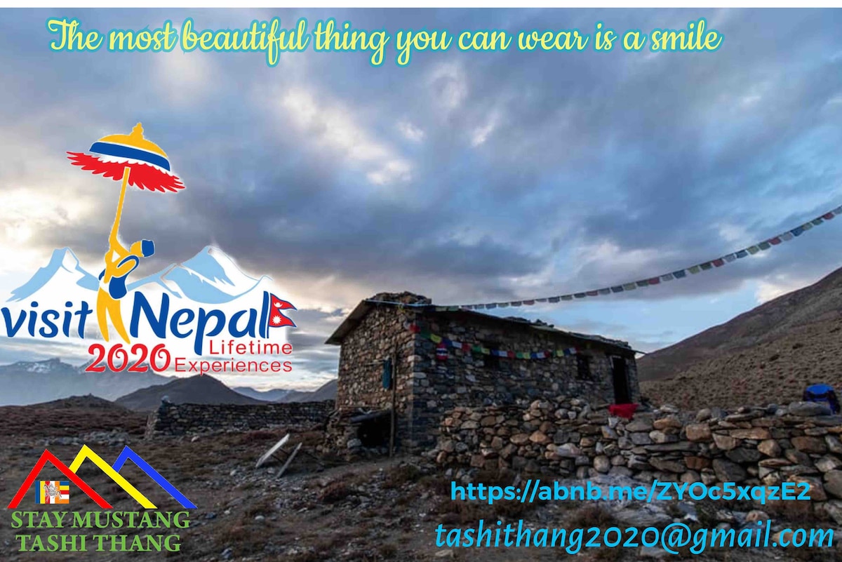 A rustic stone hut is situated against a backdrop of rolling hills and dramatic clouds. Colorful prayer flags flutter in the breeze above the hut, contributing to the serene atmosphere of the surrounding landscape.
