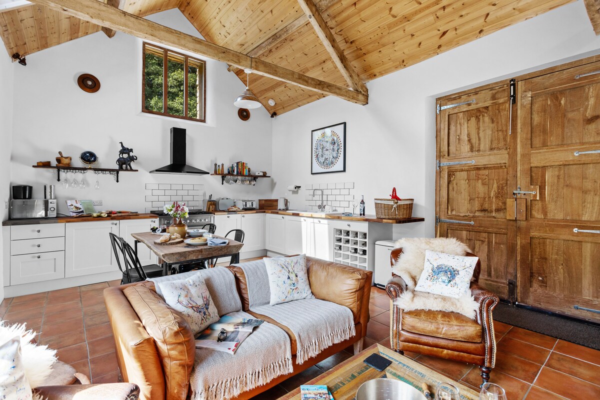 An open-plan kitchen and living area features a high wooden ceiling and rustic decor. A comfortable leather sofa is positioned opposite a wooden dining table. The kitchen is equipped with modern appliances and a spacious island, illuminated by natural light streaming through large windows.