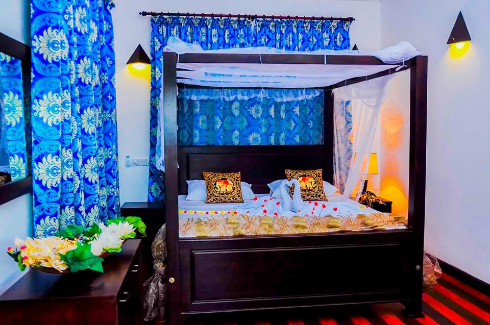 A four-poster bed is highlighted with decorative cushions and a flowing canopy. Bright blue curtains frame the window, while soft lighting emanates from wall sconces. A floral arrangement is placed beside the bed, contributing to the room's calming aesthetic.
