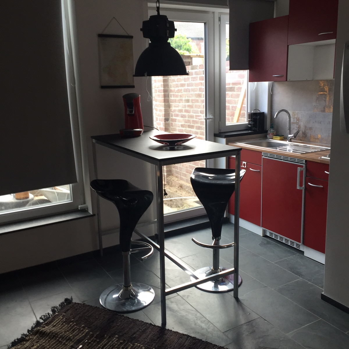 A modern kitchenette is shown, featuring sleek red cabinetry and stainless steel appliances. A small bar table with two black stools offers a space for dining. Natural light enters through the window, illuminating the minimalist design and tiled floor.