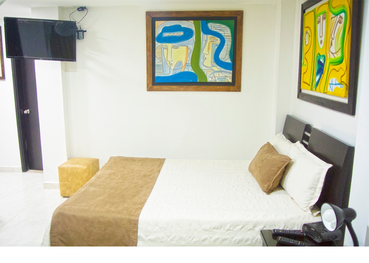 A modern room features a spacious bed adorned with neutral tones and colorful artwork above. A television is mounted on the wall, while a small decorative stool and nightstand add functionality to the space. Natural light enhances the inviting atmosphere.