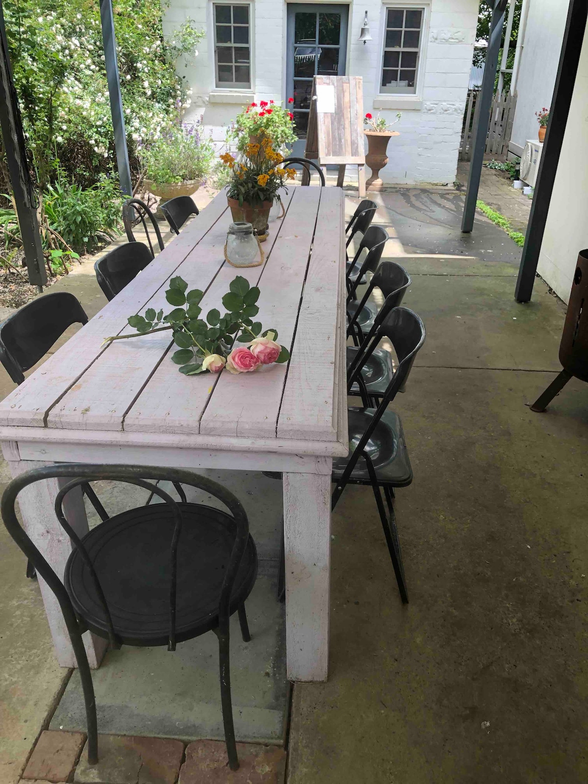 A long, rustic wooden table is surrounded by black chairs in a shaded outdoor space. A floral arrangement sits in a jar at the center of the table. The green foliage and nearby flowers create a serene garden ambiance.