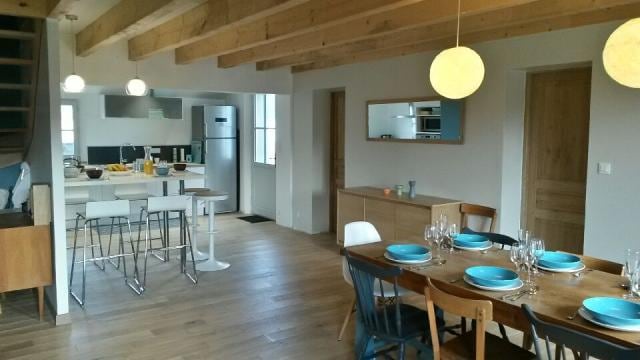 A spacious open-plan living area features a large dining table set for twelve with blue dishware. Bar stools are arranged at a kitchen island, enhancing the communal space. The high ceiling showcases wooden beams, while natural light fills the room from multiple windows.