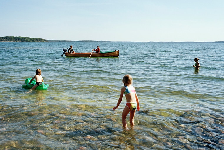 Children play in shallow water near a rocky shoreline, while two adults row a wooden boat in the background. The clear water reveals stones beneath, and the scene is set against a calm sea and a distant shoreline lined with trees.