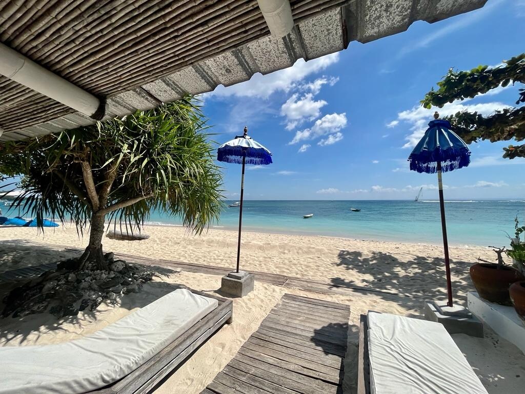 A sandy beach is visible through a shaded area featuring wooden loungers. Two blue and white umbrellas offer protection from the sun, and lush greenery frames the scene. The calm waters of the lagoon and a clear blue sky enhance the tranquil atmosphere.